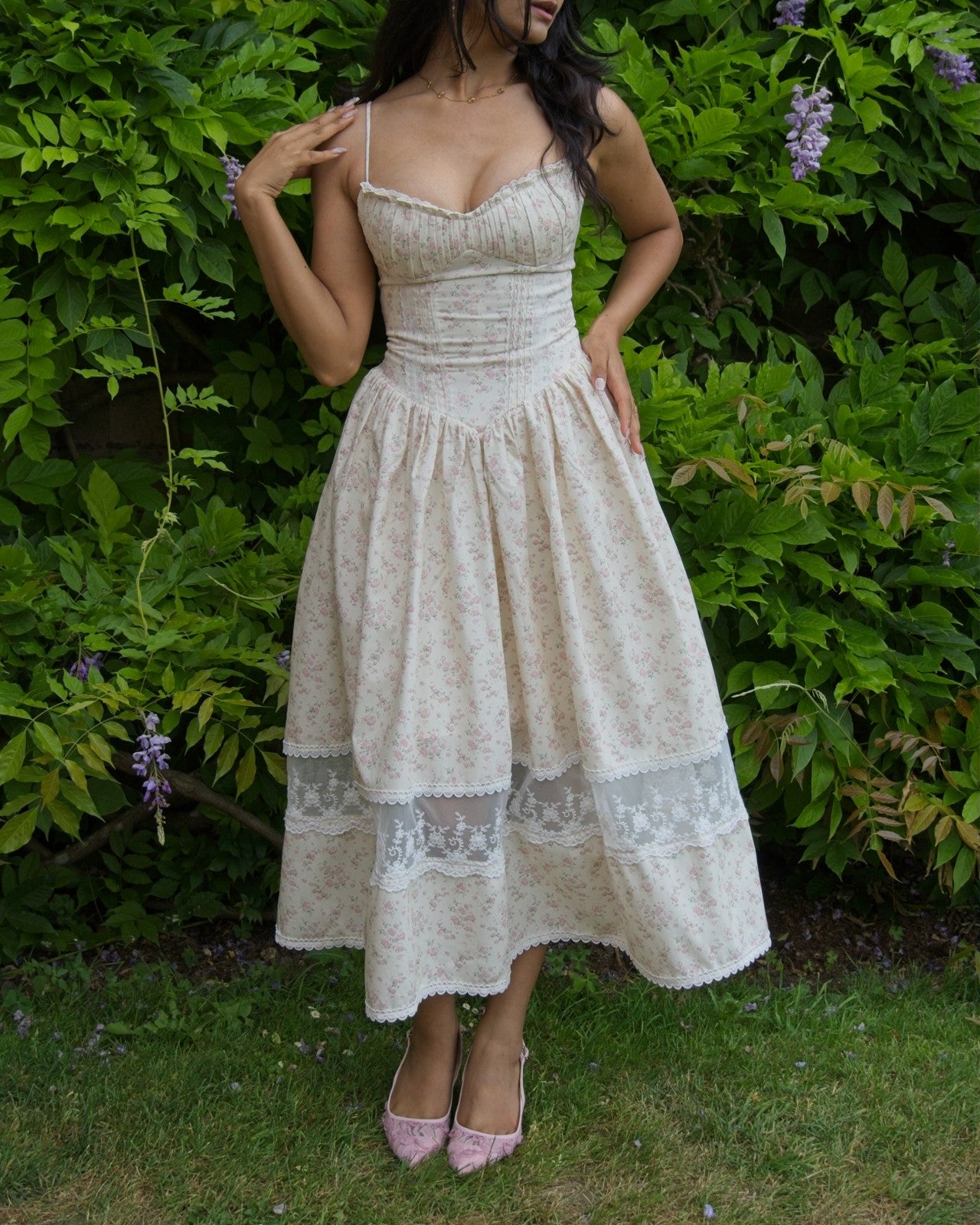 A woman wearing a cream-colored floral midi dress with lace trim and pink pointed-toe heels, standing outdoors in front of a green garden wall with purple wisteria flowers.
