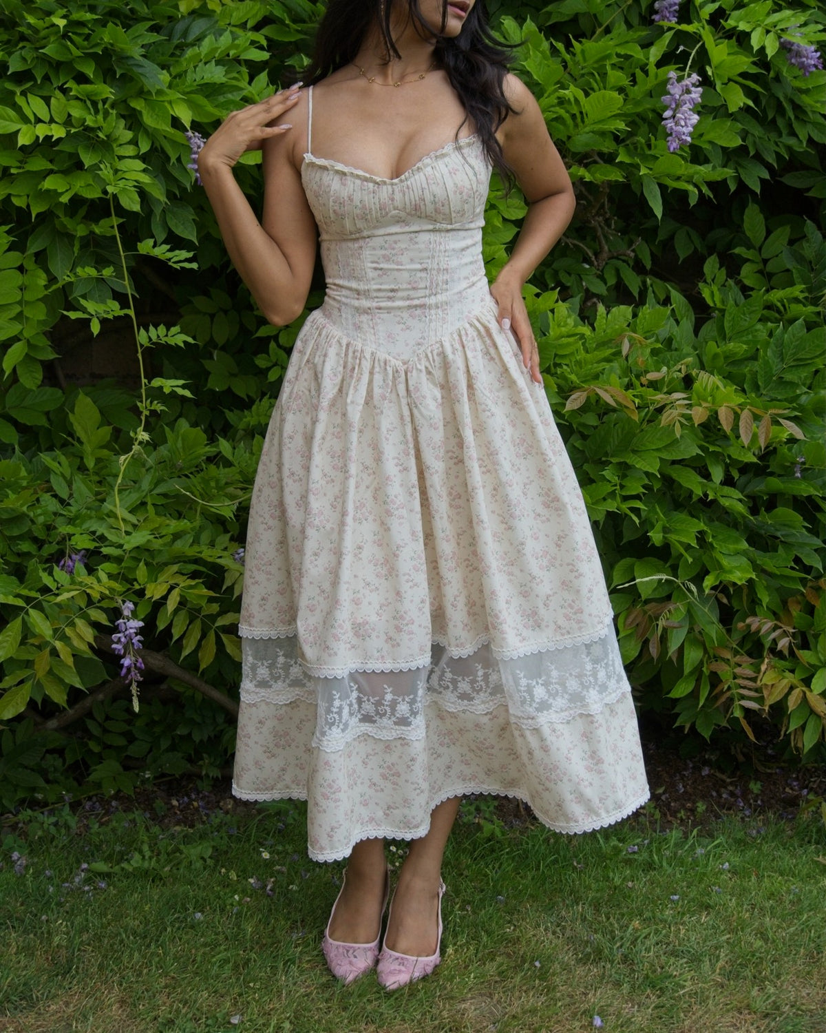 A woman wearing a cream-colored floral midi dress with lace trim and pink pointed-toe heels, standing outdoors in front of a green garden wall with purple wisteria flowers.