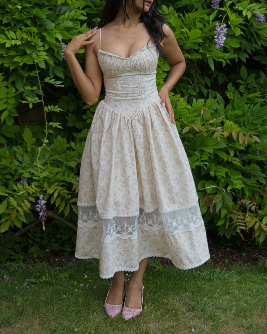 A woman wearing a cream-colored floral midi dress with lace trim and pink pointed-toe heels, standing outdoors in front of a green garden wall with purple wisteria flowers.
